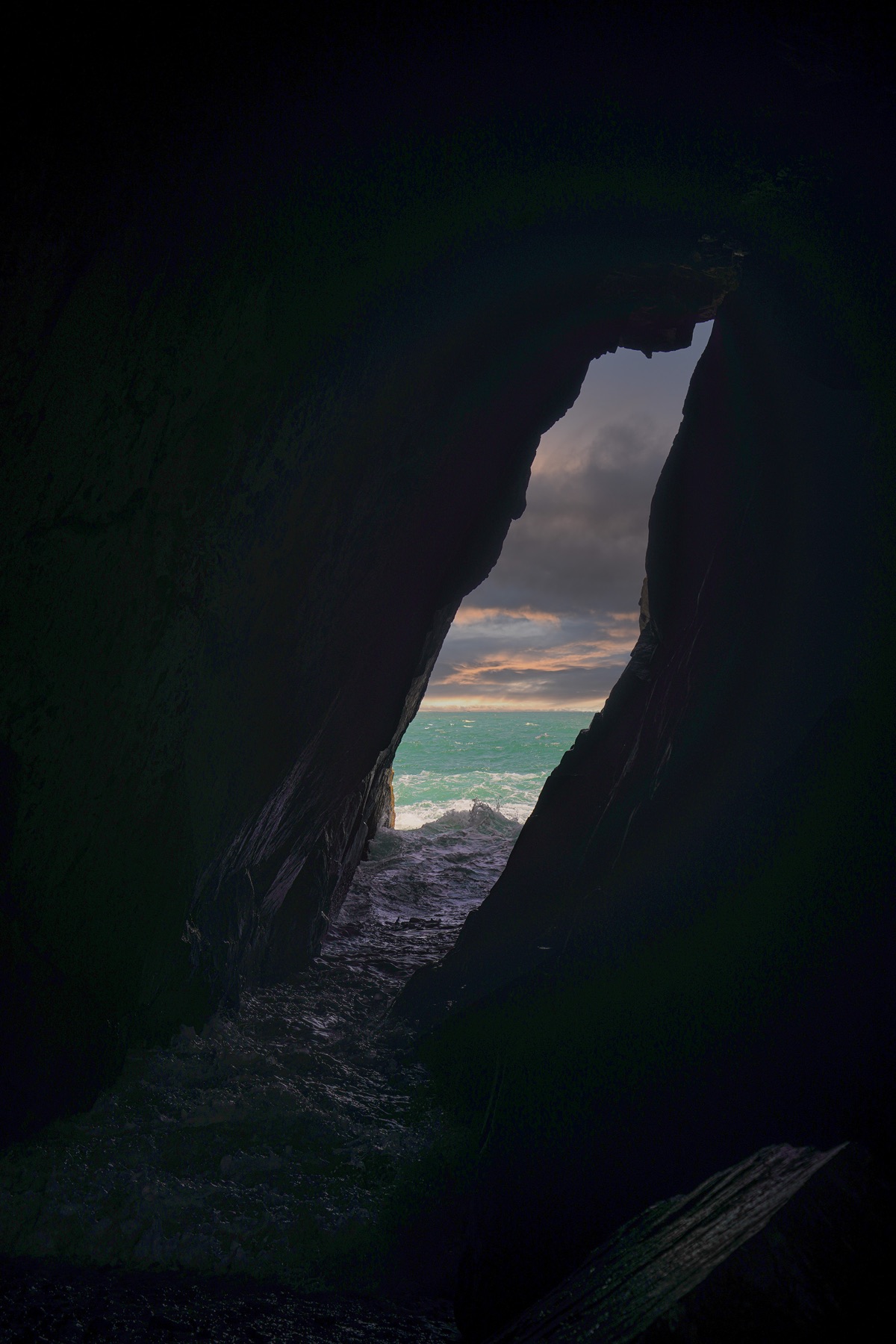 A narrow crevice in dark rock frames churning sea and sunset sky.