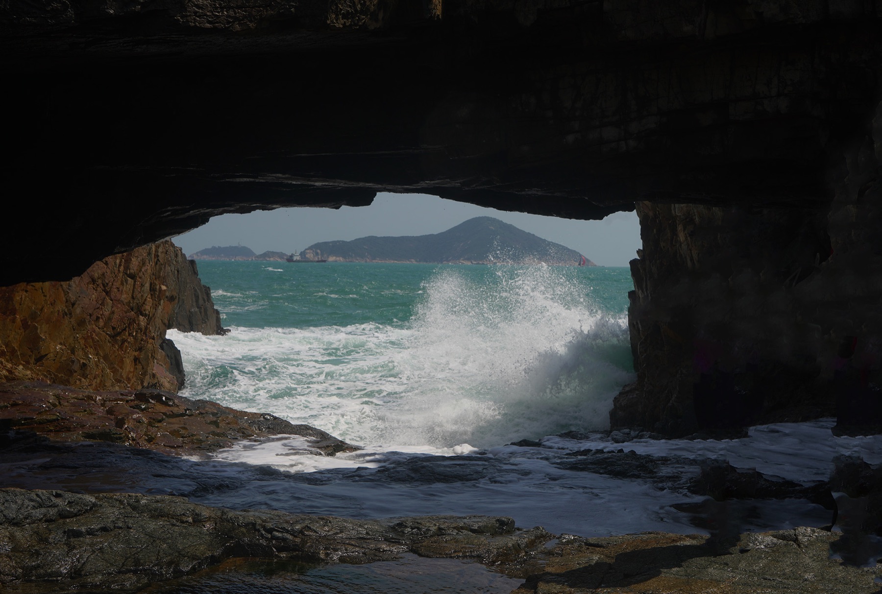Waves explode through a rocky cavern mouth, revealing a peaceful island beyond.
